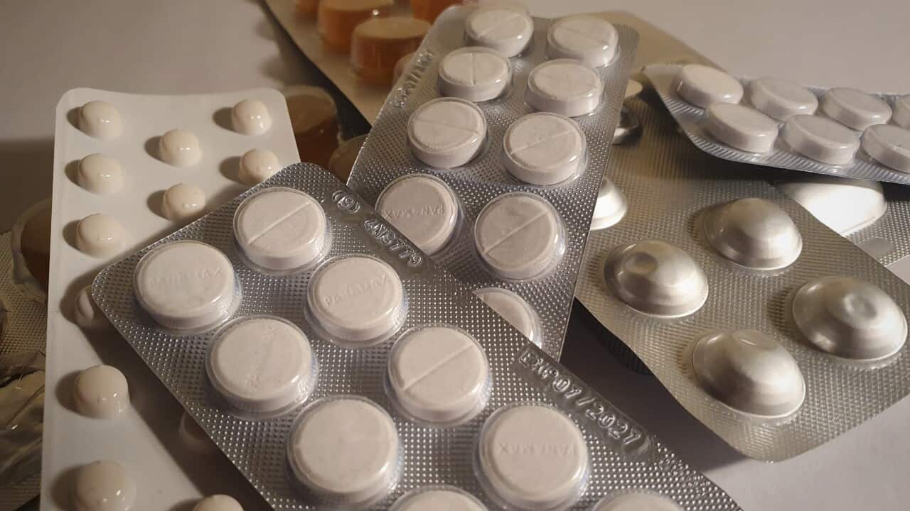 A high-angle, close-up shot shows multiple blister packs of pills scattered on a white surface. The most prominent pack in the foreground contains large, round white tablets.