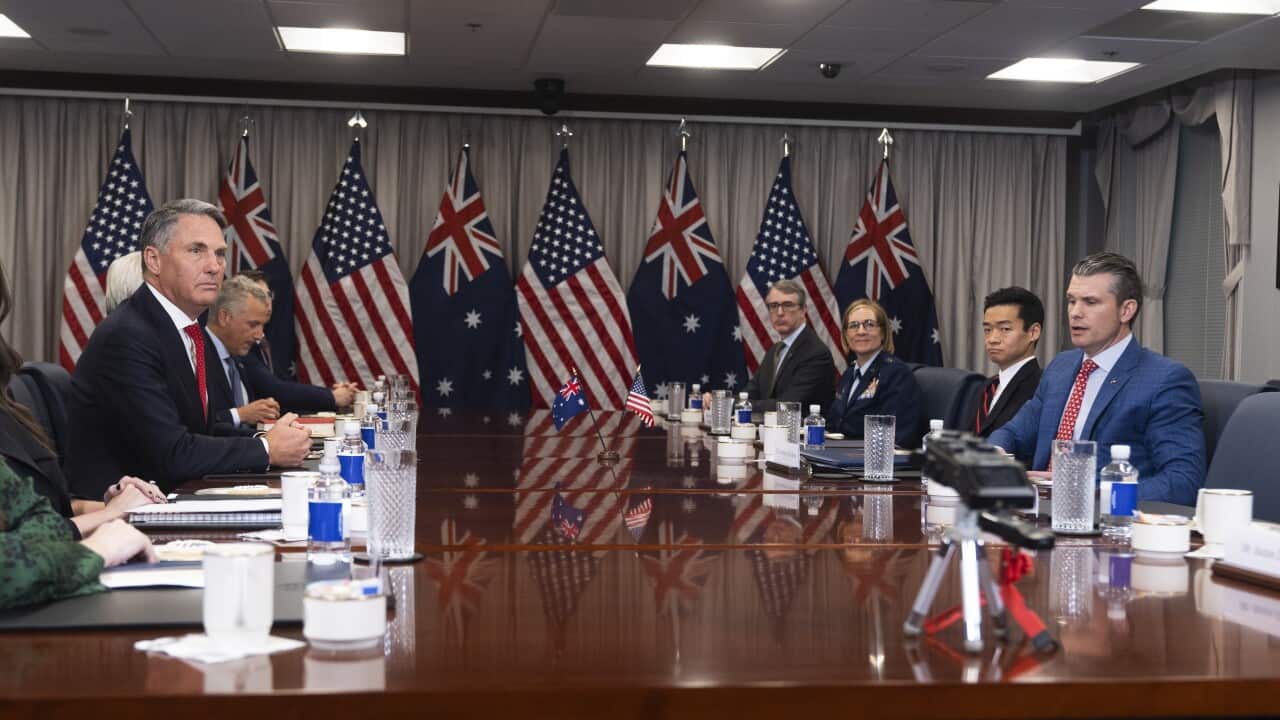 A group of people wearing formal attire sit at a wooden table. The flags of the US and Australia hang behind them.