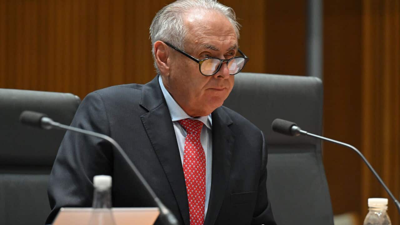An older white man wearing a dark suit and glasses, seated at a table before a microphone.