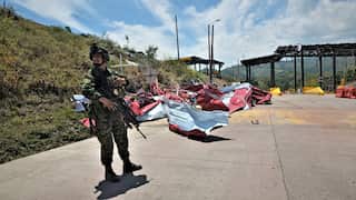 Armed soldier standing near destroyed red metal wreckage at a road tollbooth.