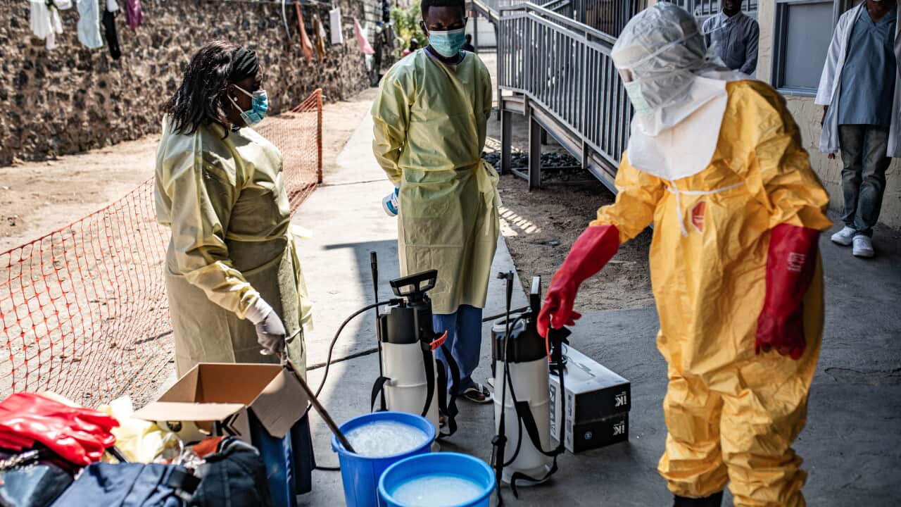 Three health workers, one in a full yellow hazmat suit, prepare decontamination sprayers and blue buckets filled with a milky liquid in an outdoor facility. All wear face masks.