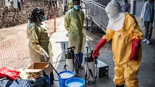Three health workers, one in a full yellow hazmat suit, prepare decontamination sprayers and blue buckets filled with a milky liquid in an outdoor facility. All wear face masks.