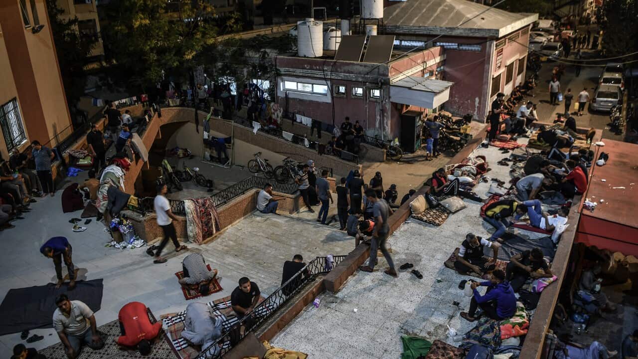 People line the entryway around a hospital, some on prayer mats, others sitting with bags beside them against walls.