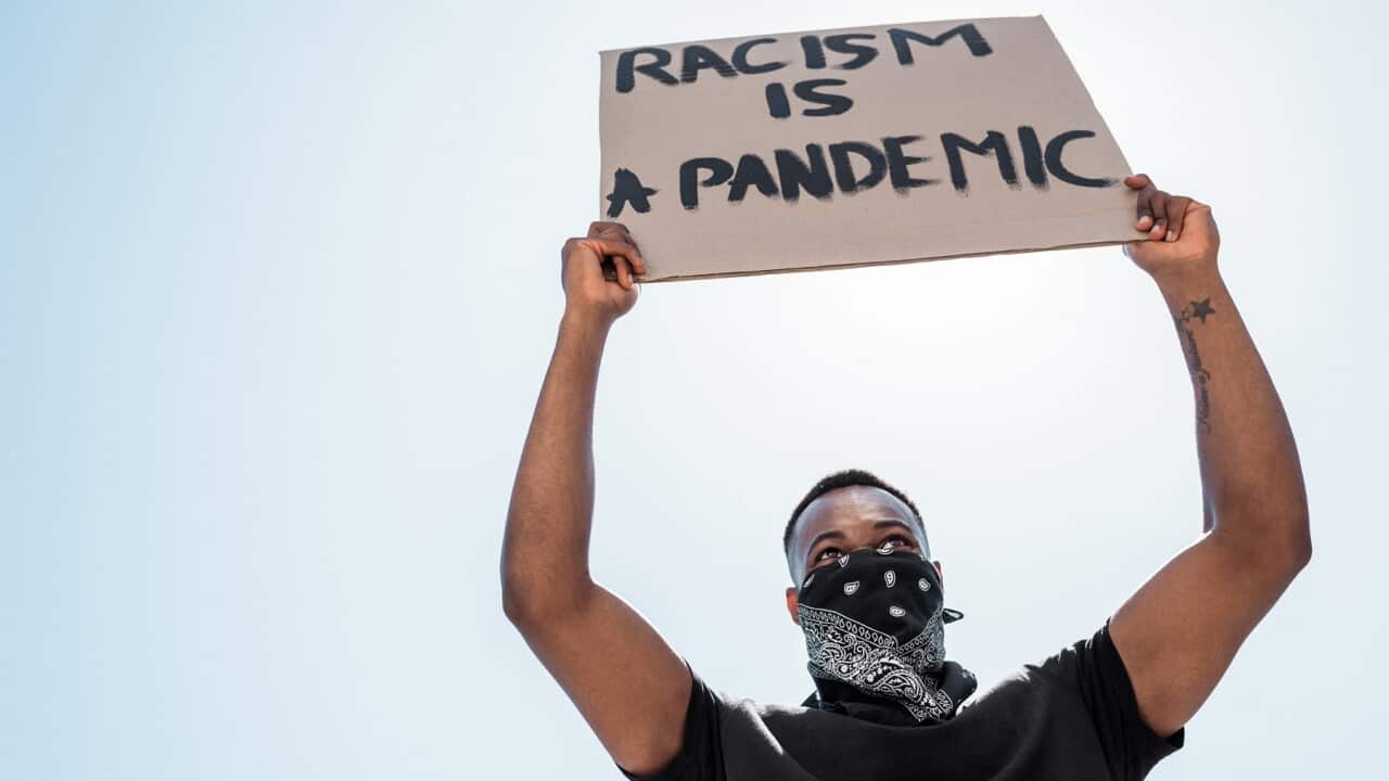 low angle view of african american man with scarf on face holding placard with racism is a pandemic lettering against blue sky