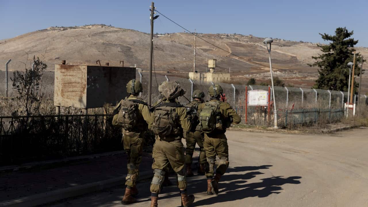A group of soldiers walks along a road in front of a fenced border.