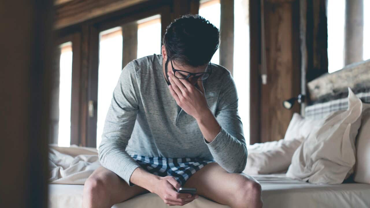Tired young man with smartphone sitting on bed at home squinting
