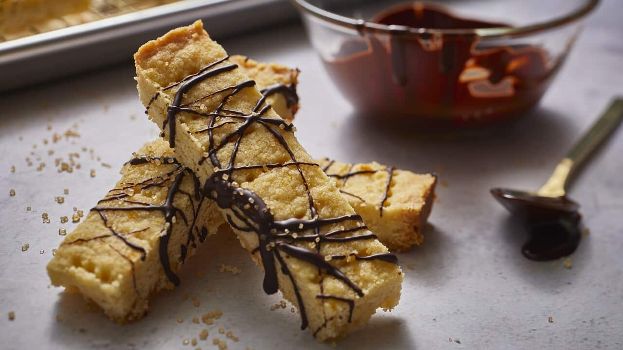 Three shortbread fingers, crisscrossed with set drizzled chocolate, sit in a pile on a bench. A tray with more bars can be seen in the background.