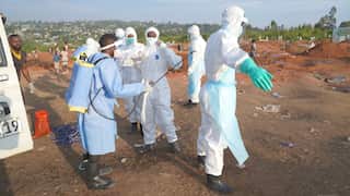 A group of people in white hazardous material suits are sprayed down by a man in blue protective clothing.