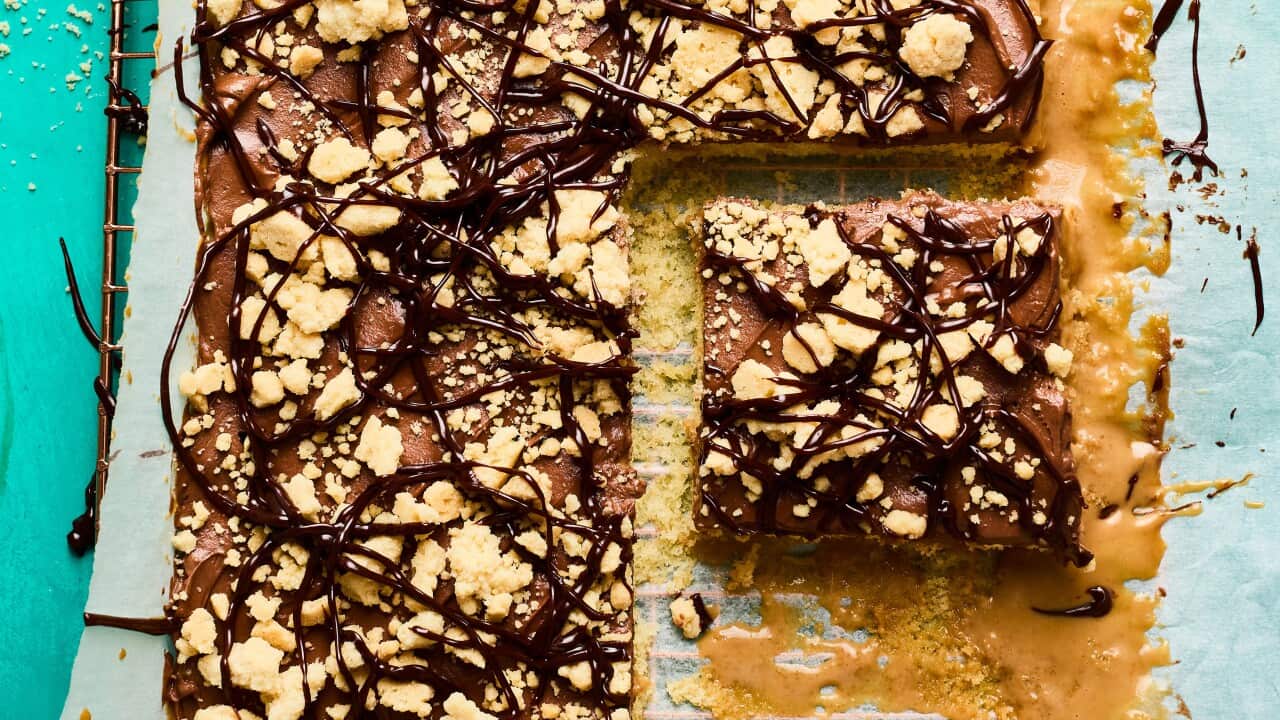 A crumble and chocolate-topped low cake sits on baking paper on a wire rack. One piece has been removed, leaving a drizzle of caramel sauce on the paper.