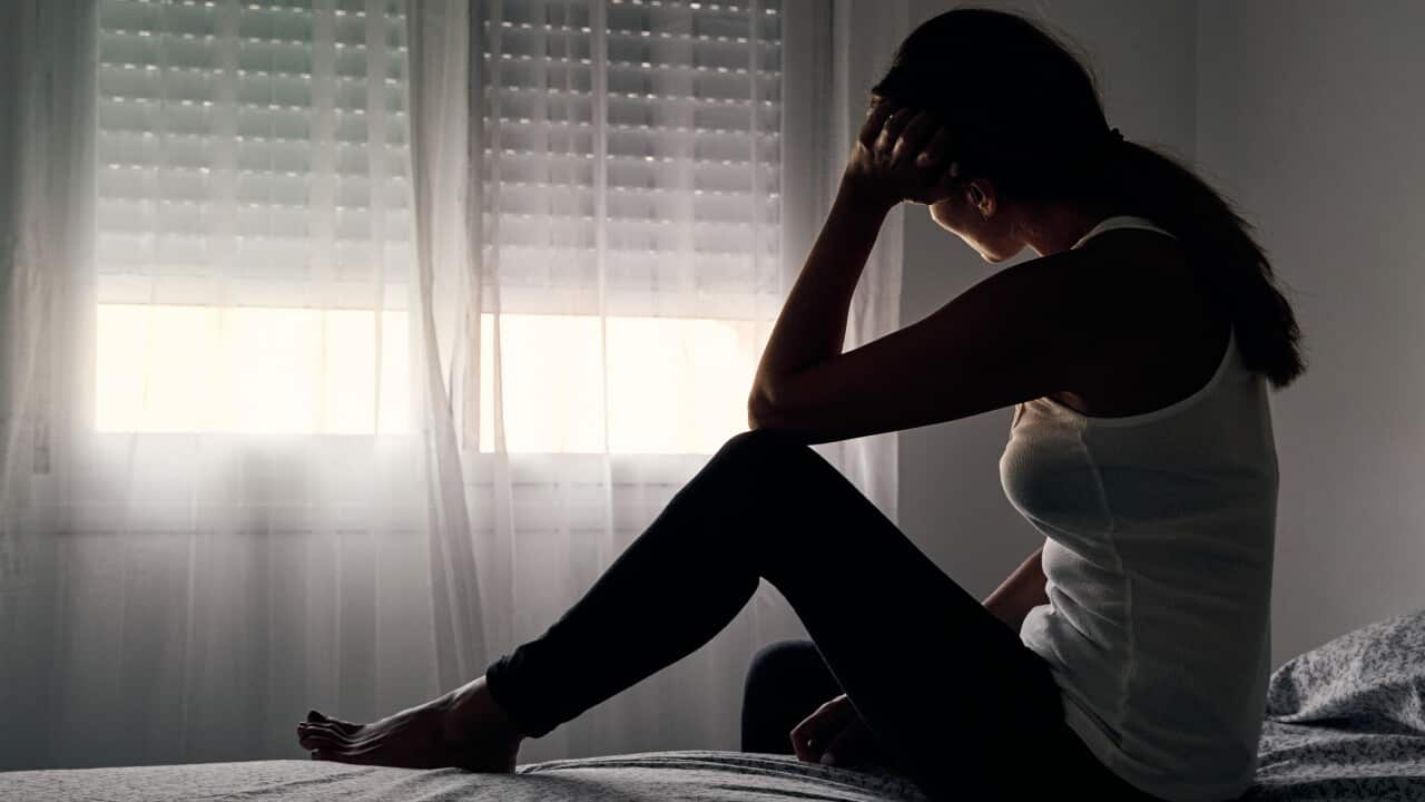 A silhouette of a woman sitting on the edge of a bed in a dimly lit room, resting her head on her hand in front of a window with closed blinds.