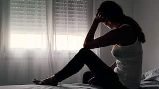 A silhouette of a woman sitting on the edge of a bed in a dimly lit room, resting her head on her hand in front of a window with closed blinds.