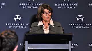 A middle aged white woman talks at a blakc lectern with microphones toward her while a succession of reporters look on.