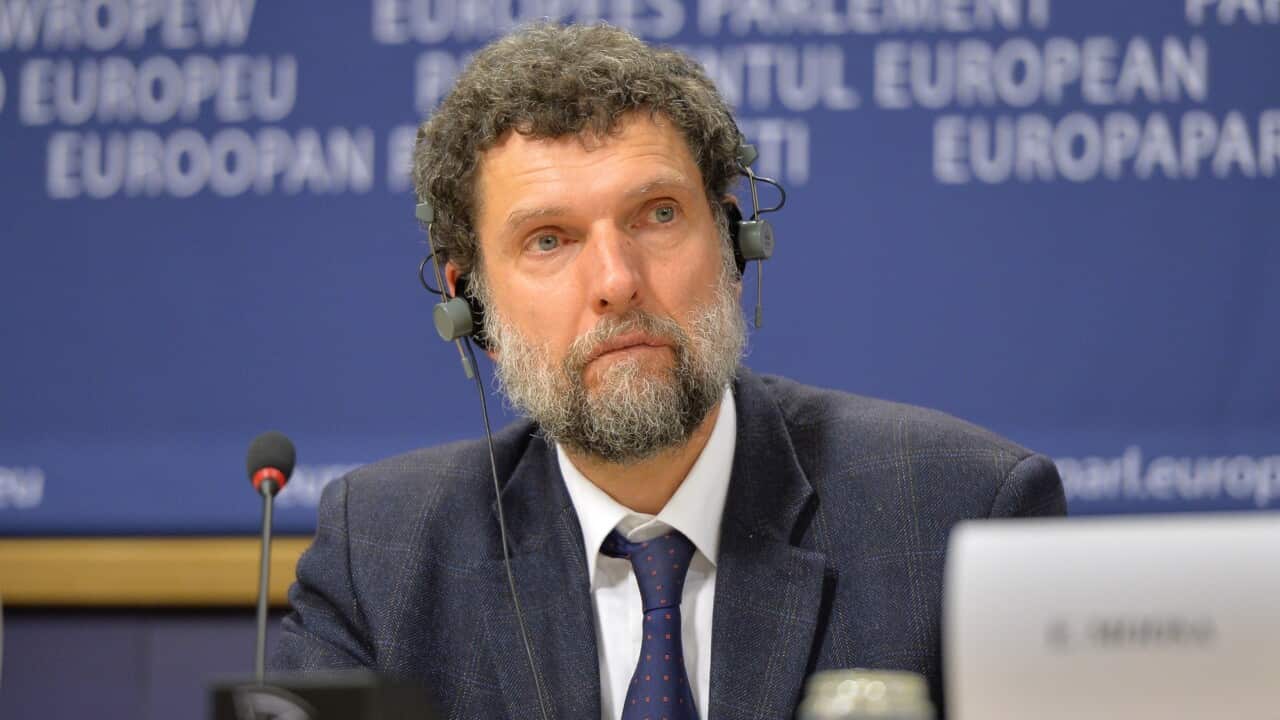 Osman Kavala sits at a a podium wearing a headset and suit.