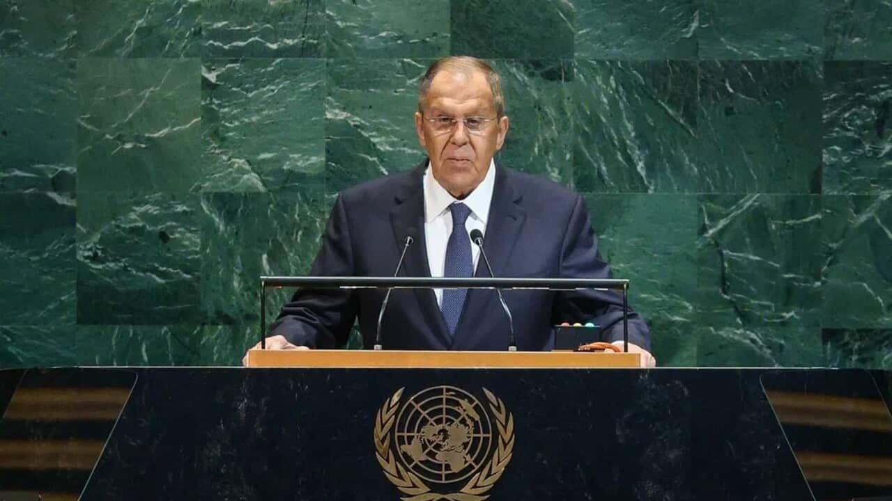 A close-up photograph shows Russian Foreign Minister Sergei Lavrov speaking from a podium at the United Nations General Assembly. He is a bald man wearing dark-rimmed glasses and a navy suit with a blue patterned tie, standing against a backdrop of mottled green stone.