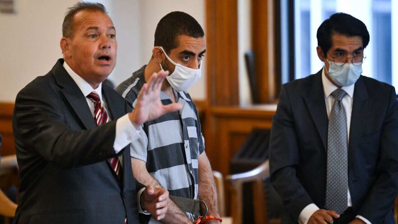 A man in handcuffs, a blue-striped prison jumpsuit, and a white face mask stands between two men in suits