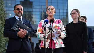 Three people, a man and two women, stand before a pair of microphones outdoors. The man and woman on the left and right are dressed in dark suits while the woman in the centre is dressed in a colourful blazer and eyeglasses.