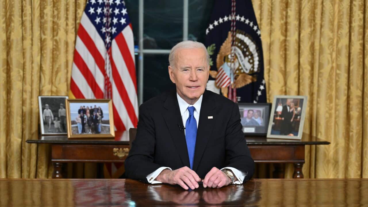 Joe Biden, in a dark suit and blue tie, sits at a large wooden table with the US flag and US coat of arms flag behind him.
