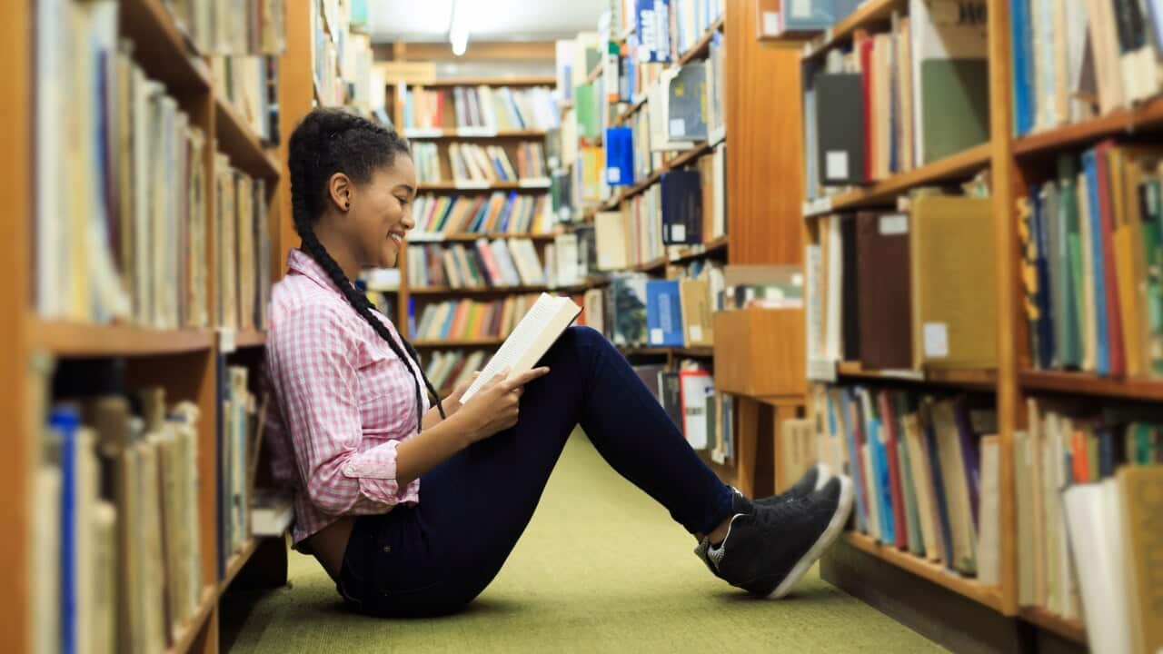 A teenage girl sitting on the floor of a library smiling while reading a book.