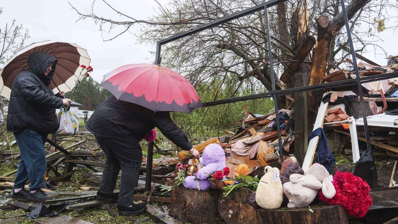 Two women holding umbrellas put flowers by the rubble of a destroyed house.