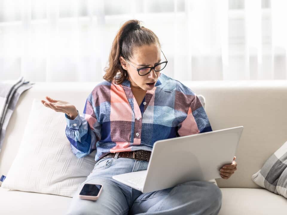 A woman stares at her laptop with her mouth open and a blank expression, throwing her hand up in surprise. Her frustrated reaction and smartphone resting on her thigh suggest that she has just encountered an unexpected problem or bad news.