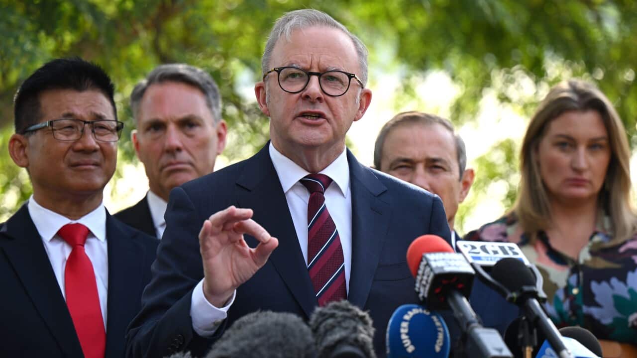 Anthony Albanese gestures while speaking in front of several media-branded microphone. He is flanked by Richard Marles, Roger Cook and several others.