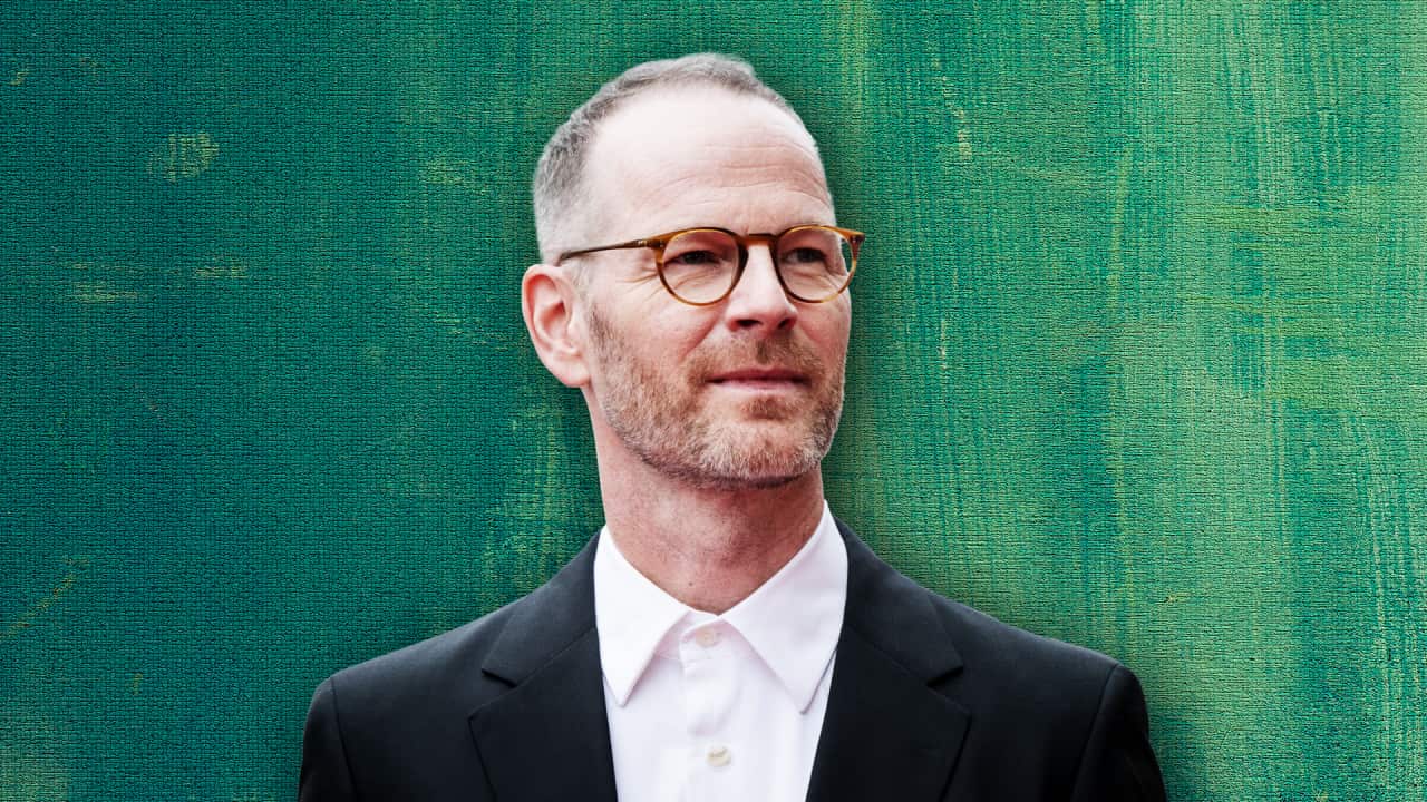 A man in a black suit and glasses looks sideways as he stands in front of a green background.