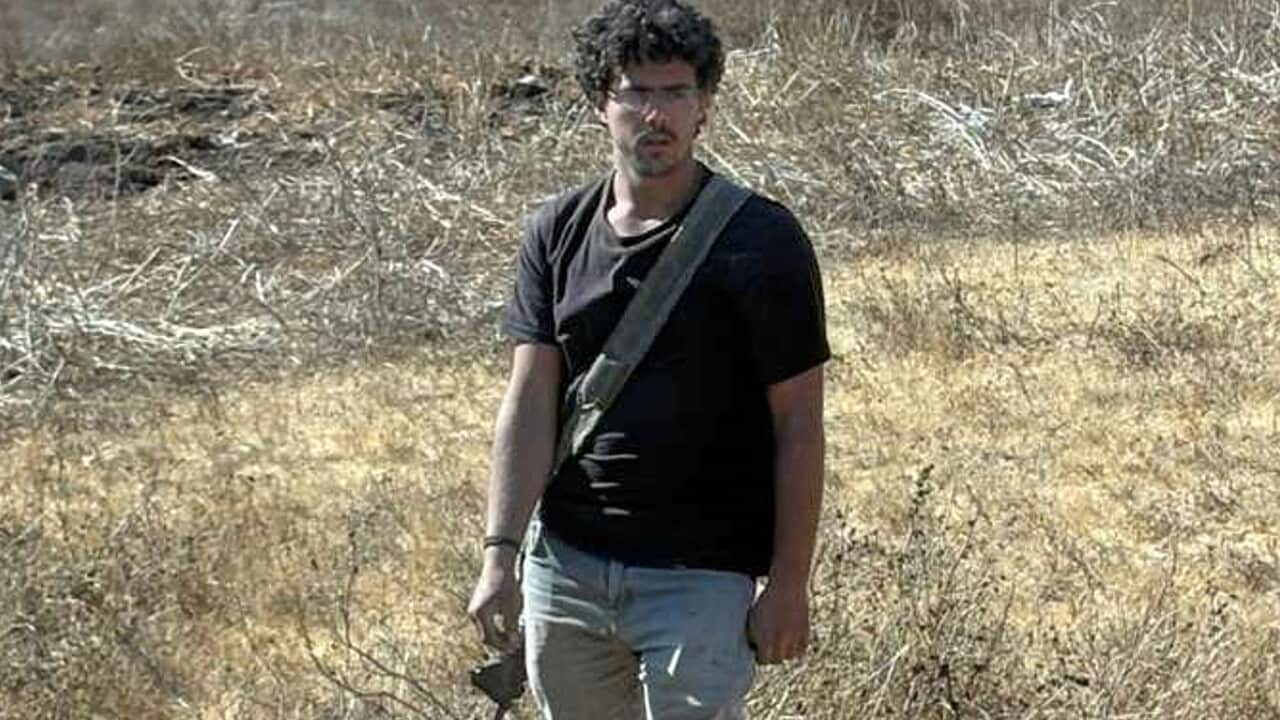 A full-length shot of a man with dark curly hair and glasses, wearing a black T-shirt and jeans, standing in a dry, grassy field while carrying a rifle.