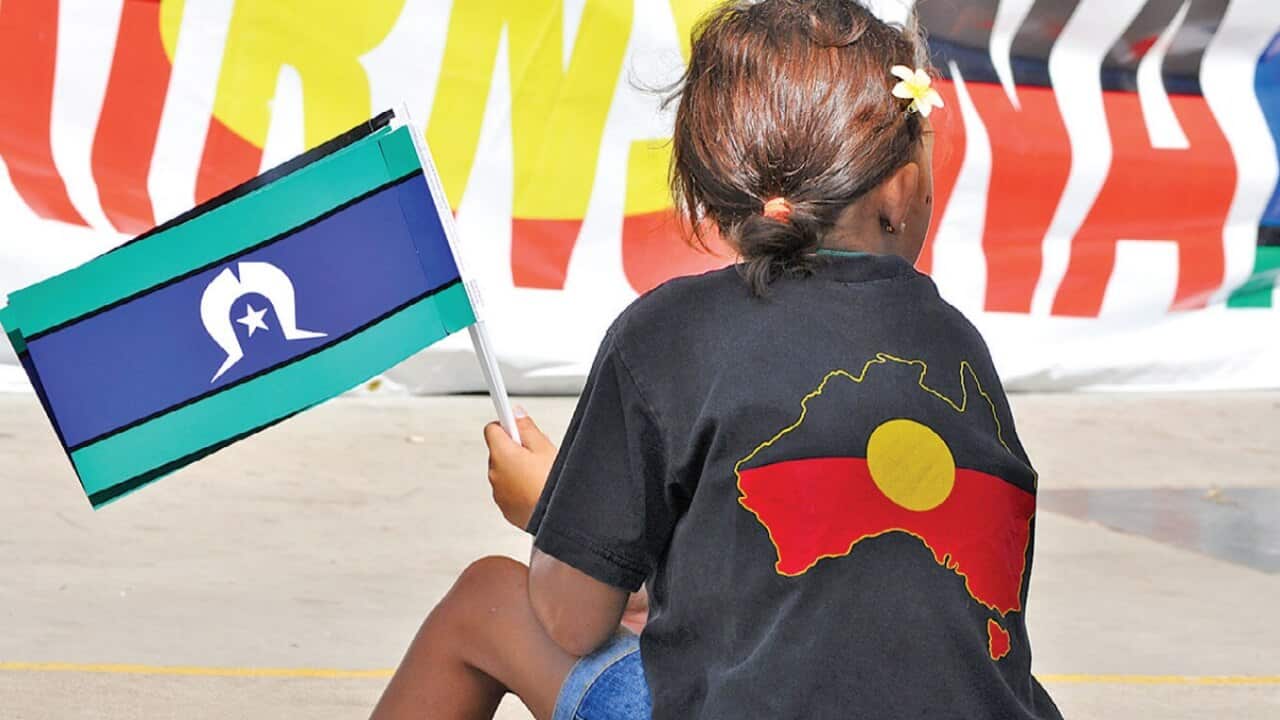 A young girl sitting down wearing a t-shirt with a Koori Flag design, holding a Torres Straits Flag
