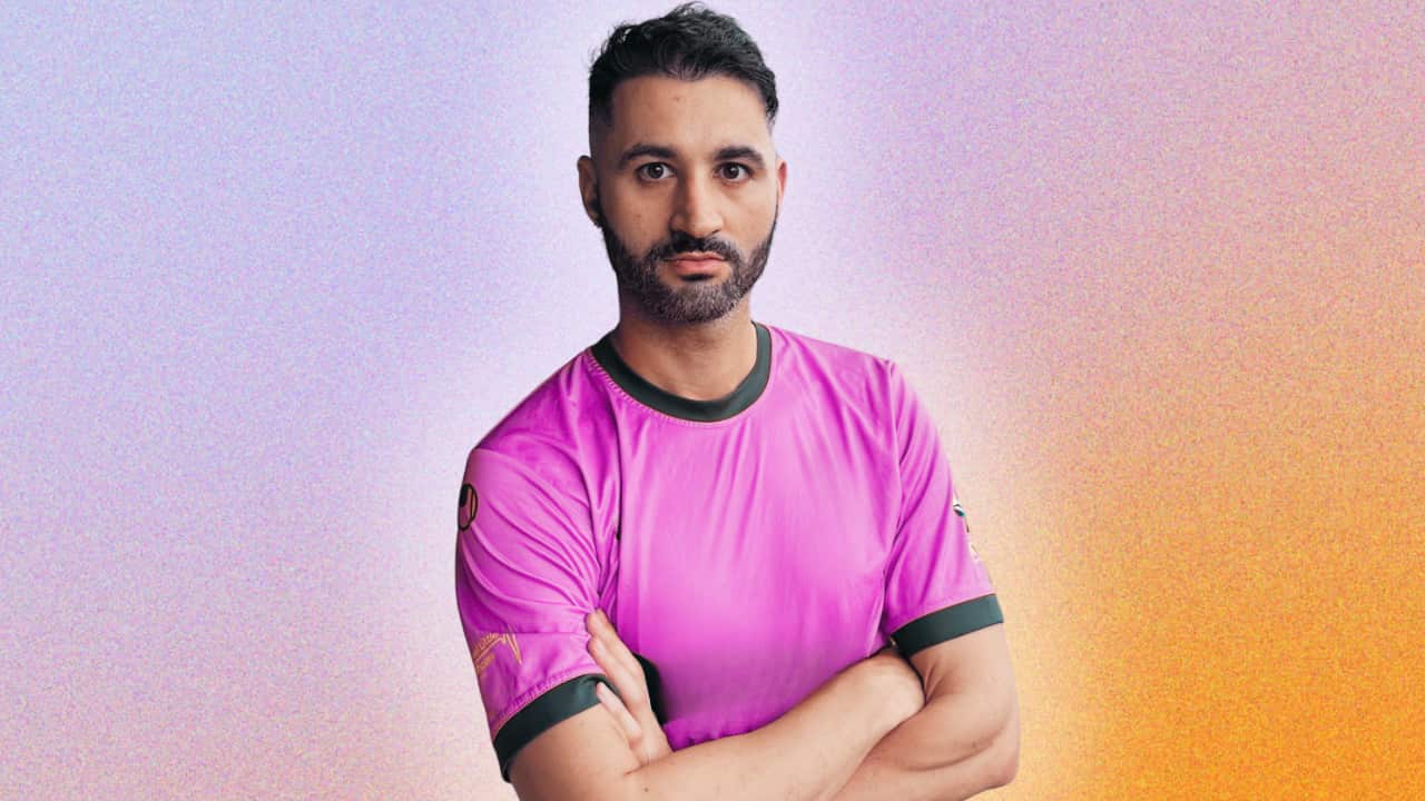A man wearing an pink football shirt has his arms crossed with a gradient of yellow and pink in the background