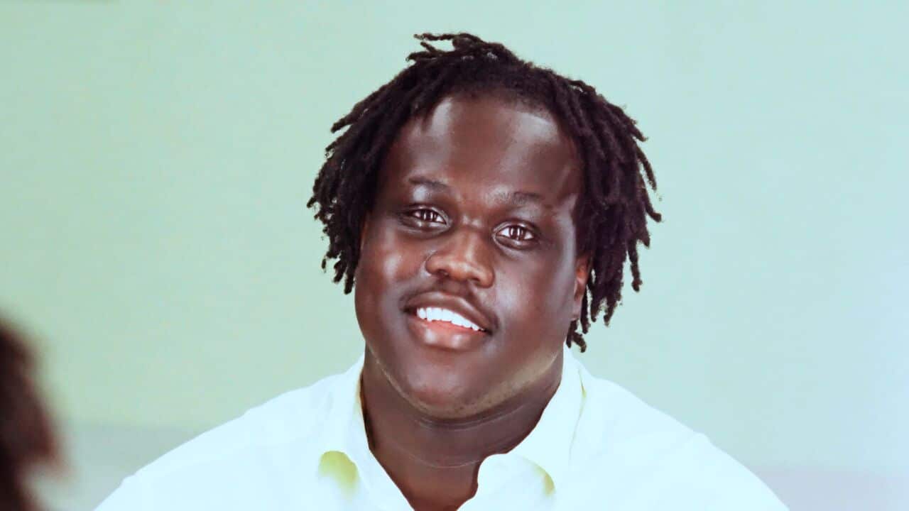 A close-up shot of a young man with his hair in short tight twists and in a white collared shirt smiling