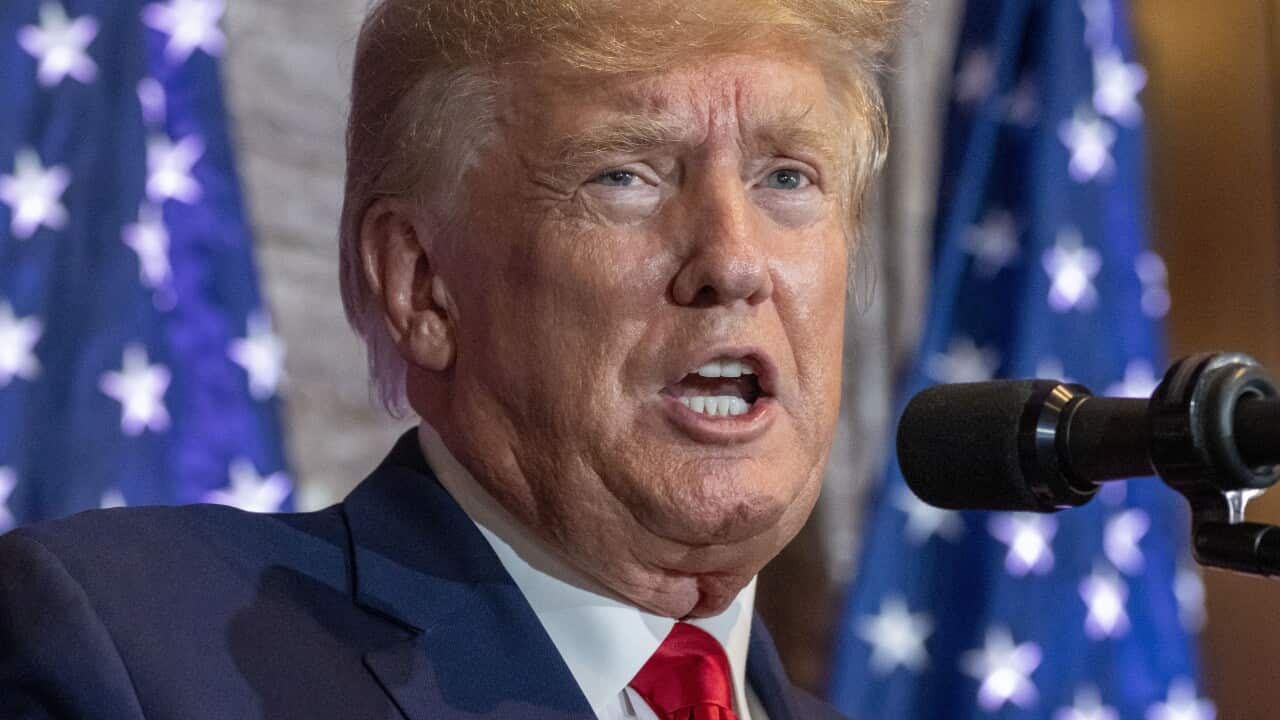 Donald Trump talks into a microphone. He is wearing a red tie and dark blue jacket.