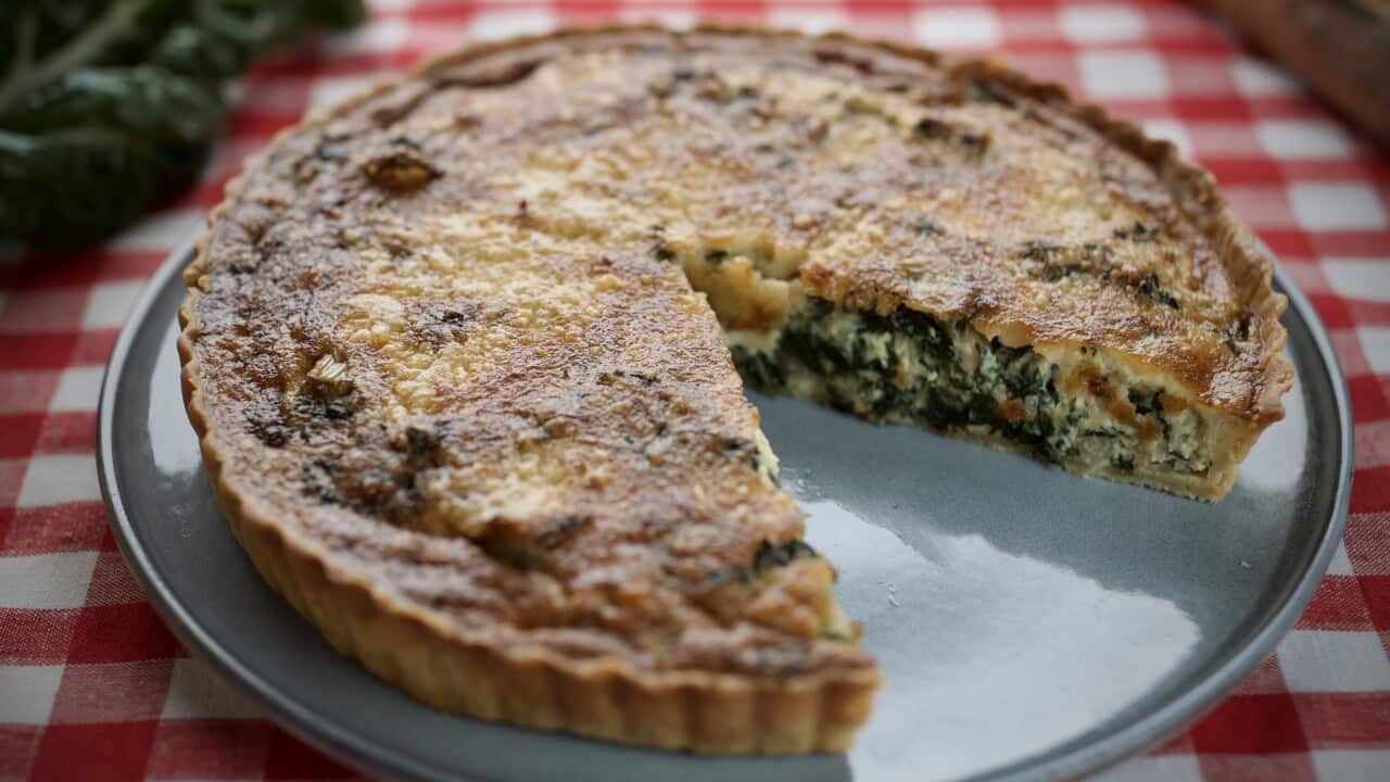 A round, shallow tart with a fluted pastry edge sits on a round plate. A large slice has been removed, showing the egg and silverbeet filling.