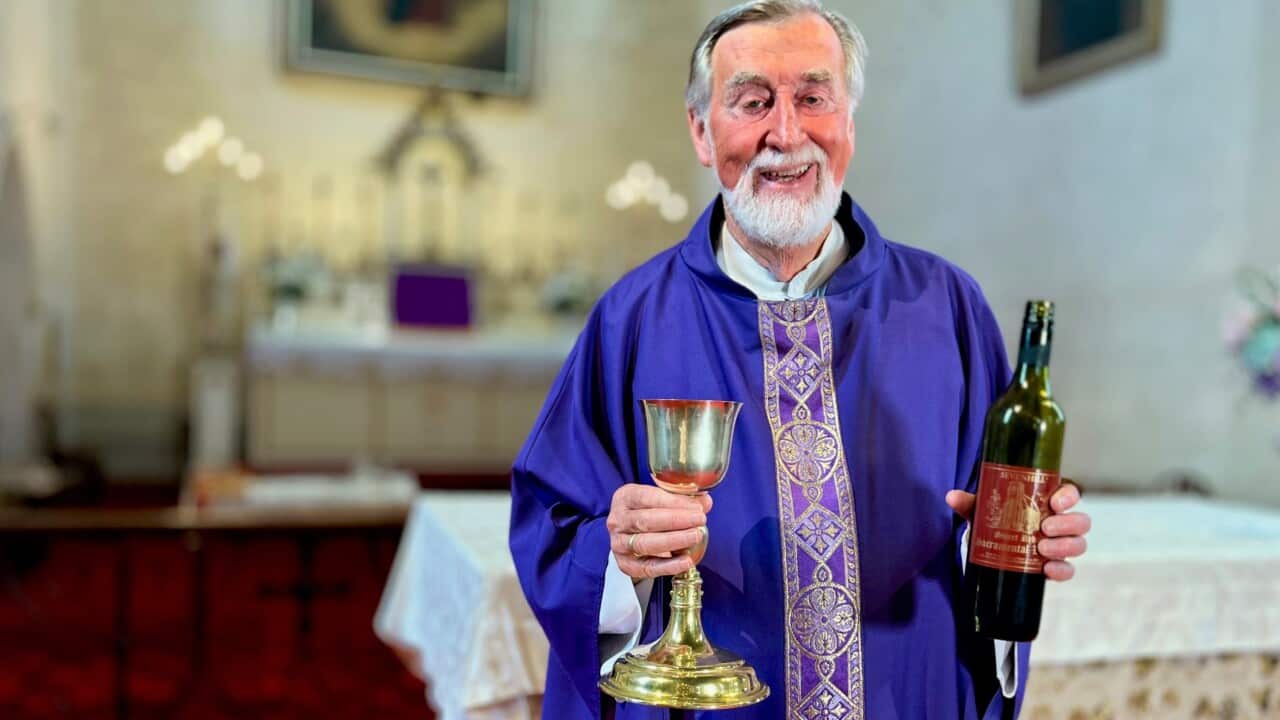 A man with a beard and wearing a purple robe stands inside a church holding a bottle of wine and a brass chalice.