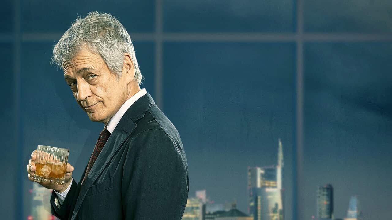 A grey-haired man in a suit, holding a whisky glass, stands in front of a window and a night skyline. He looks quizzically to one side.