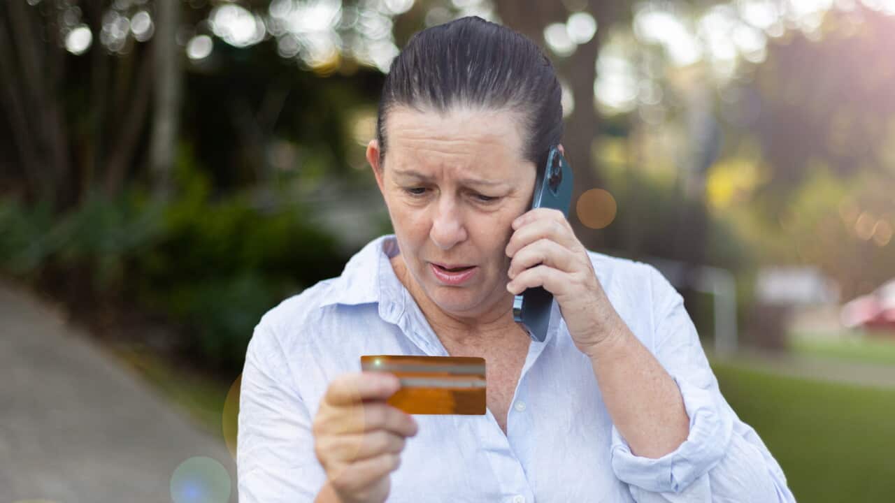 Middle aged caucasian woman looking at credit card while on the phone looking distressed and upset, wearing semi casual clothing sitting in backyard of home