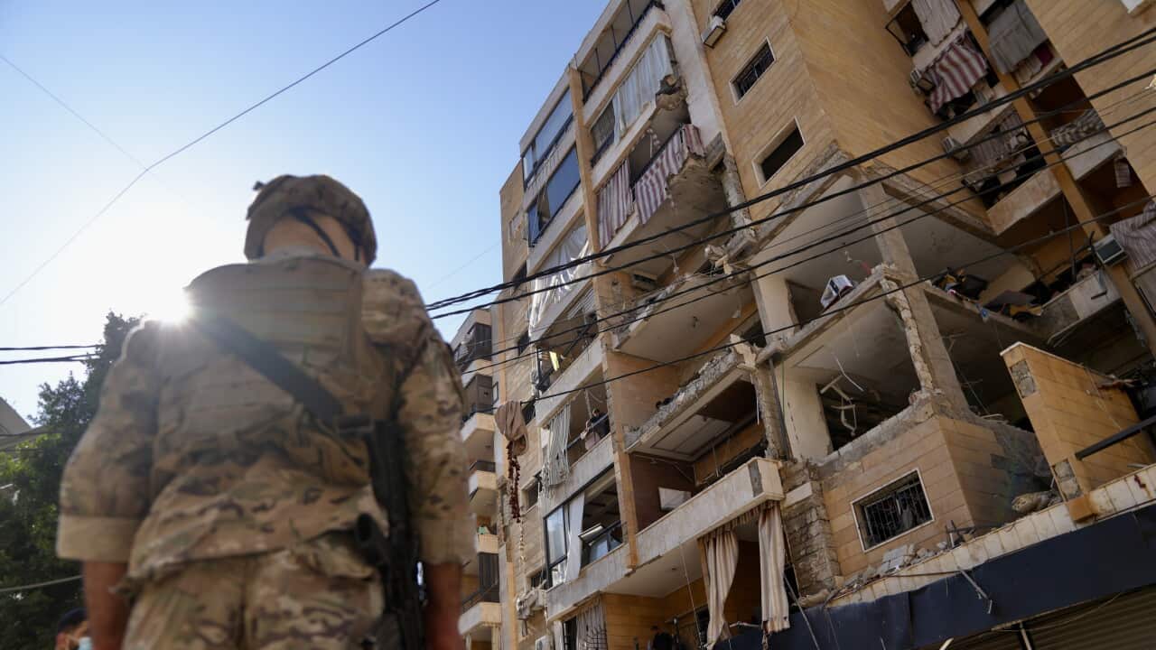 An armed soldier stands in front of a ruined bricked building.