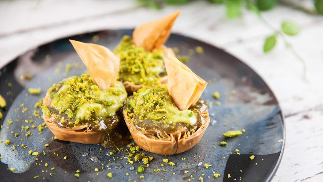 Three small filo tartlets, topped with a pale green cream and crushed pistachios, each with a pastry triangle as decoration, sit on a wide dark pottery plate.