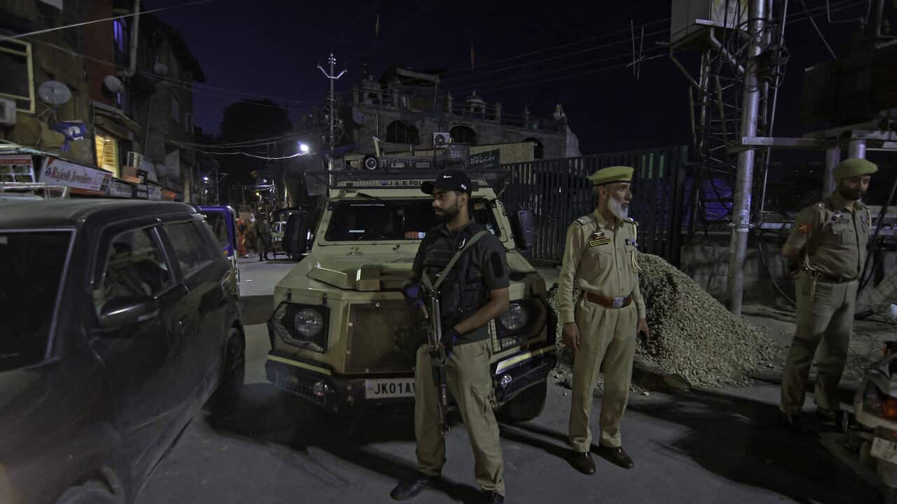 Three men dressed in camouflage clothing stand in front of a military vehicle, one holding a gun.