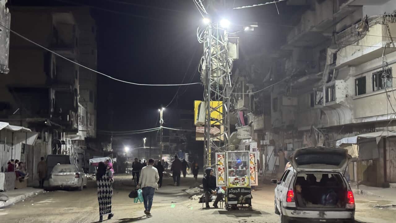 View of a residential street at night, illuminated by streetlights. A car is parked in the middle of the road with its boot open, while people walk around.