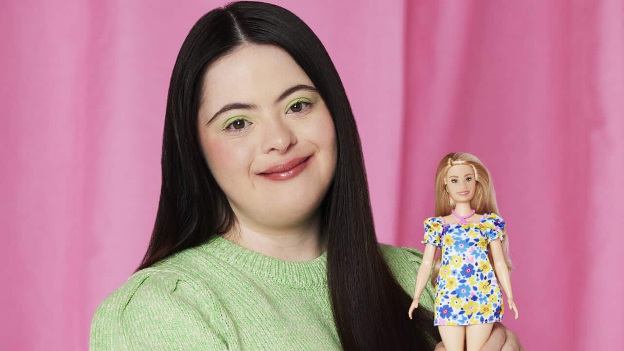 A girl in a green shirt carries a Barbie doll in front of a pink backdrop