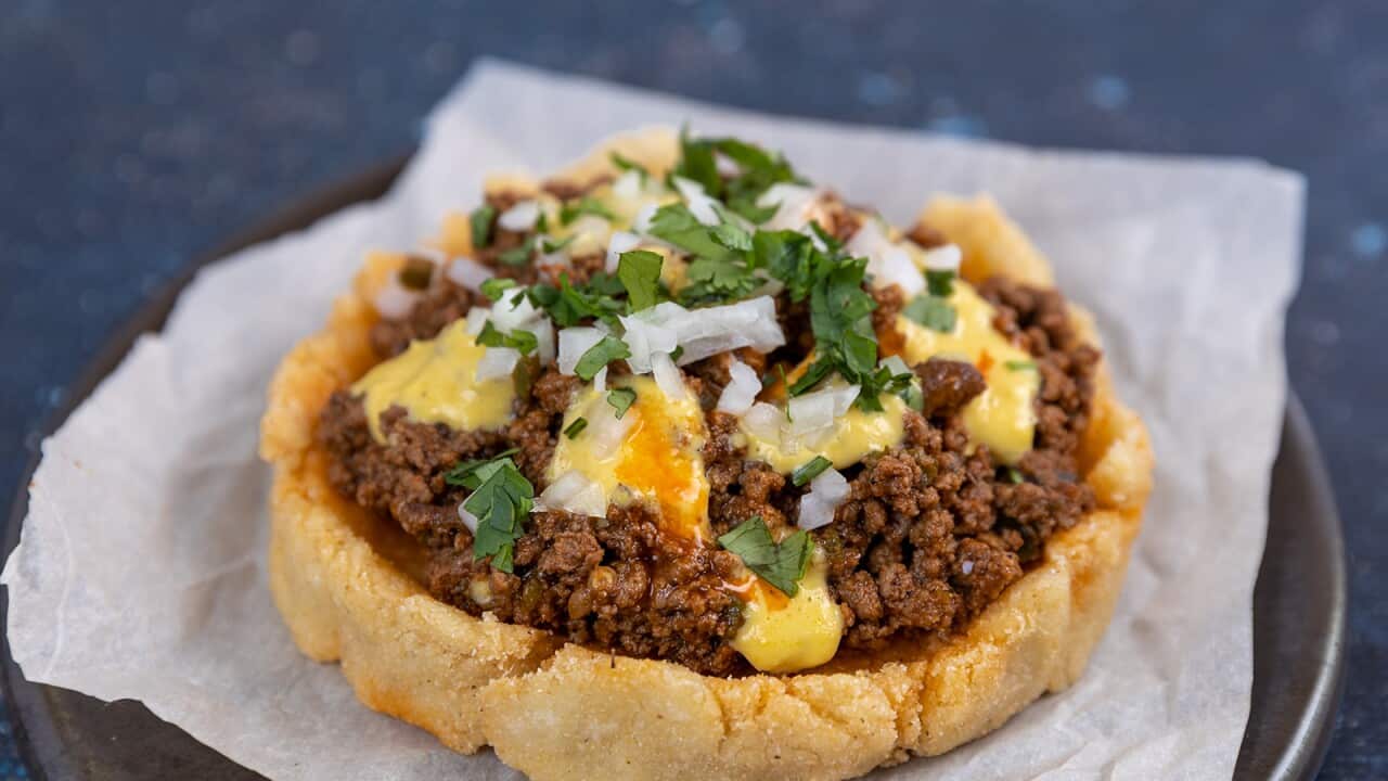 A round corn cake sits on a piece of paper, on a brown plate. The cake is topped with mince and small dollops of yellow sauce, and garnished with chopped onion and coriander.