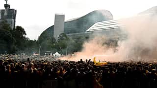 A densely packed crowd of football fans stands behind barriers at an outdoor live site, with orange smoke drifting above the audience. Many people are wearing Australia's green and gold colours, while inflatable kangaroo toys are held above the crowd. The curved W Hotel building and a high-rise under construction are visible in the background.