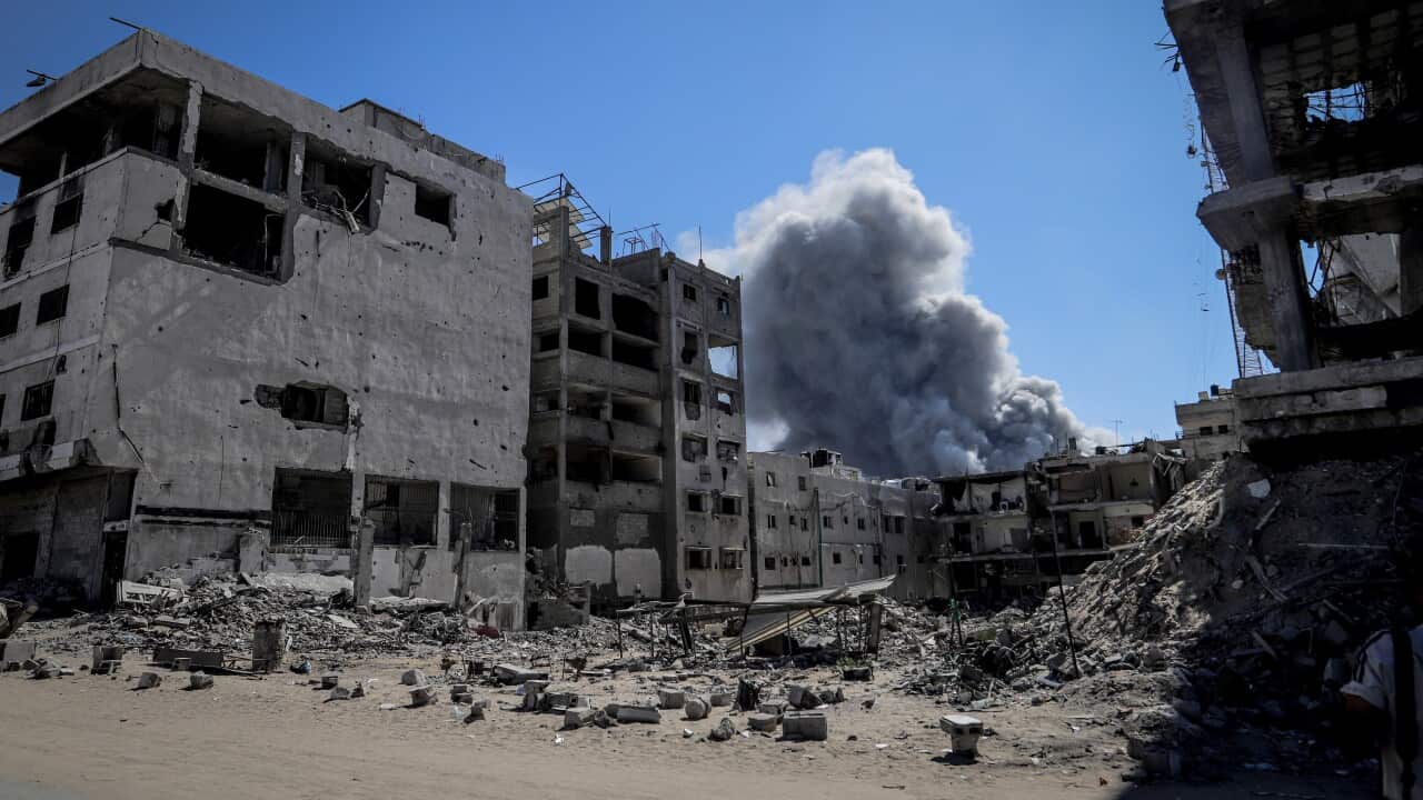 Heavily damaged and rubble-strewn buildings sit under a clear blue sky, with a large column of dark gray smoke billowing up from the centre. The surrounding structures are skeletal and pockmarked.
