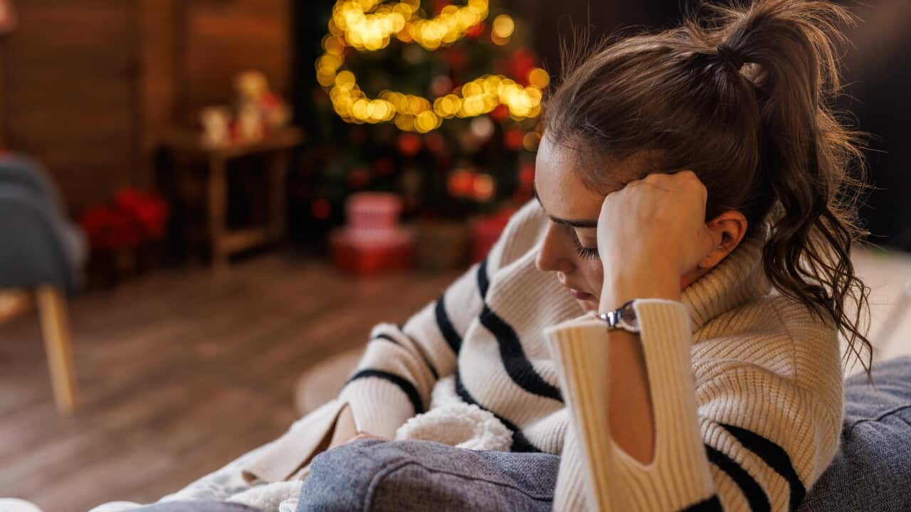 A young woman sits on a couch, her hand on her head, with a sad expression on her face.