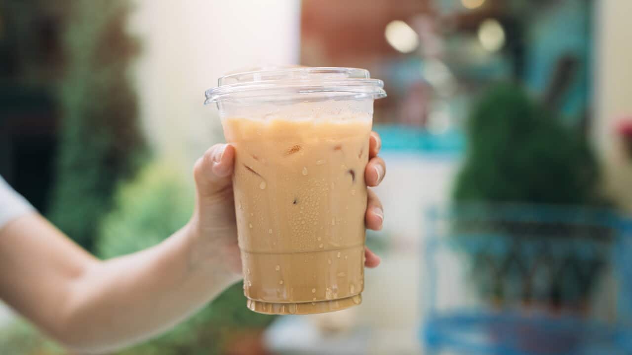A person holds a refreshing iced coffee in a clear cup, showcasing condensation on the surface. The background features soft light and greenery.
