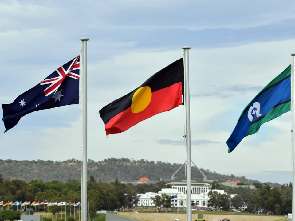 Australian flag, Aboriginal flag and Torres Strait Island flag flying on flag poles with Old Parliament House and Parliament House in Canberra in the background.