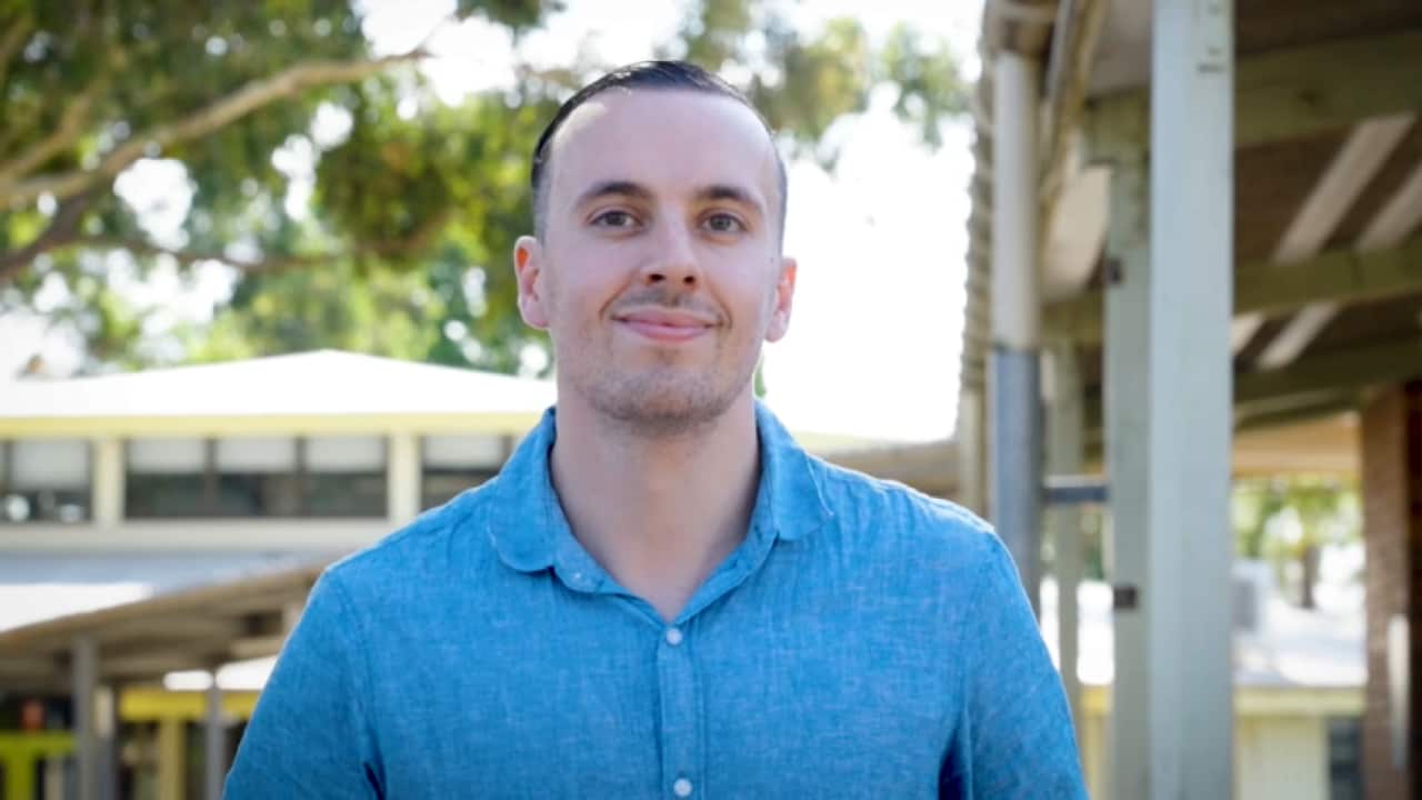 A man in a blue shirt smiles at the camera. There is a school in the background.