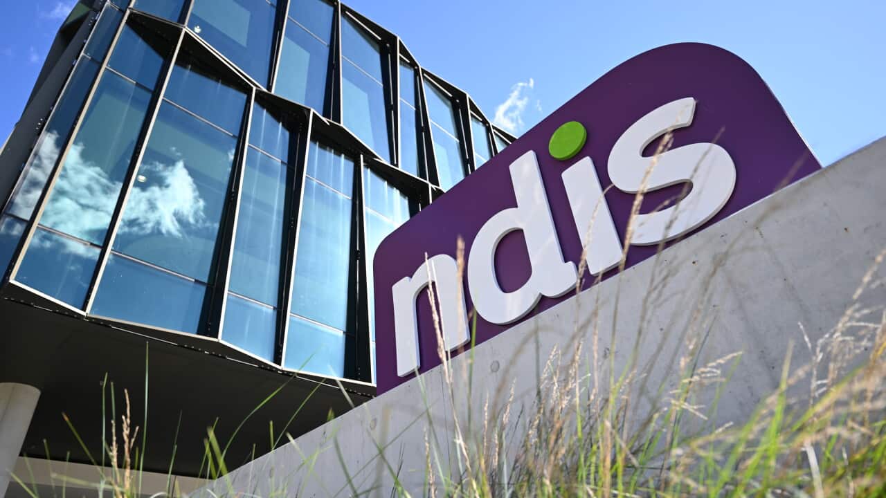 A purple sign sits in high grass outside a large building.
