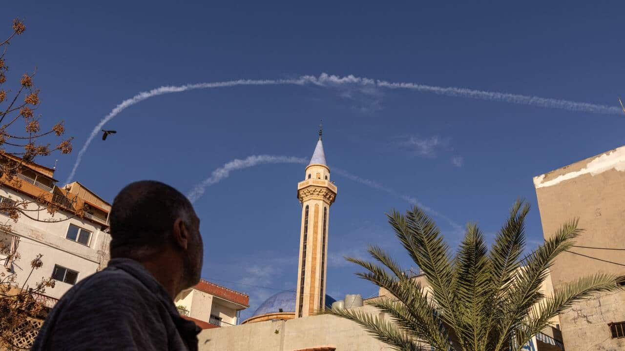 An image of Israeli airstrikes in Tyre, Lebanon. An older man looks to the sky above a parapet where two contrails from Israeli fighter jets hang in the air.