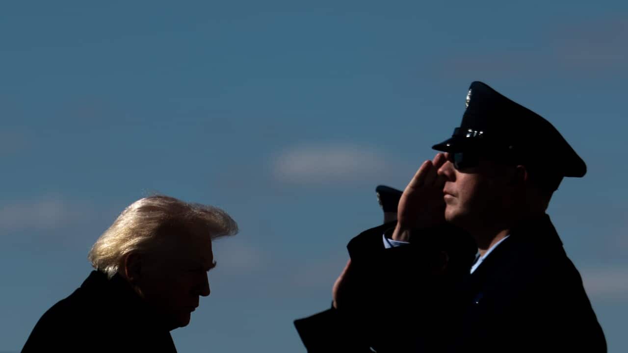US President Donald Trump boards passes a pair of military officers. The military officers faces have sunlight on them, while Trump is in shadow but for the hair on the back of his head.